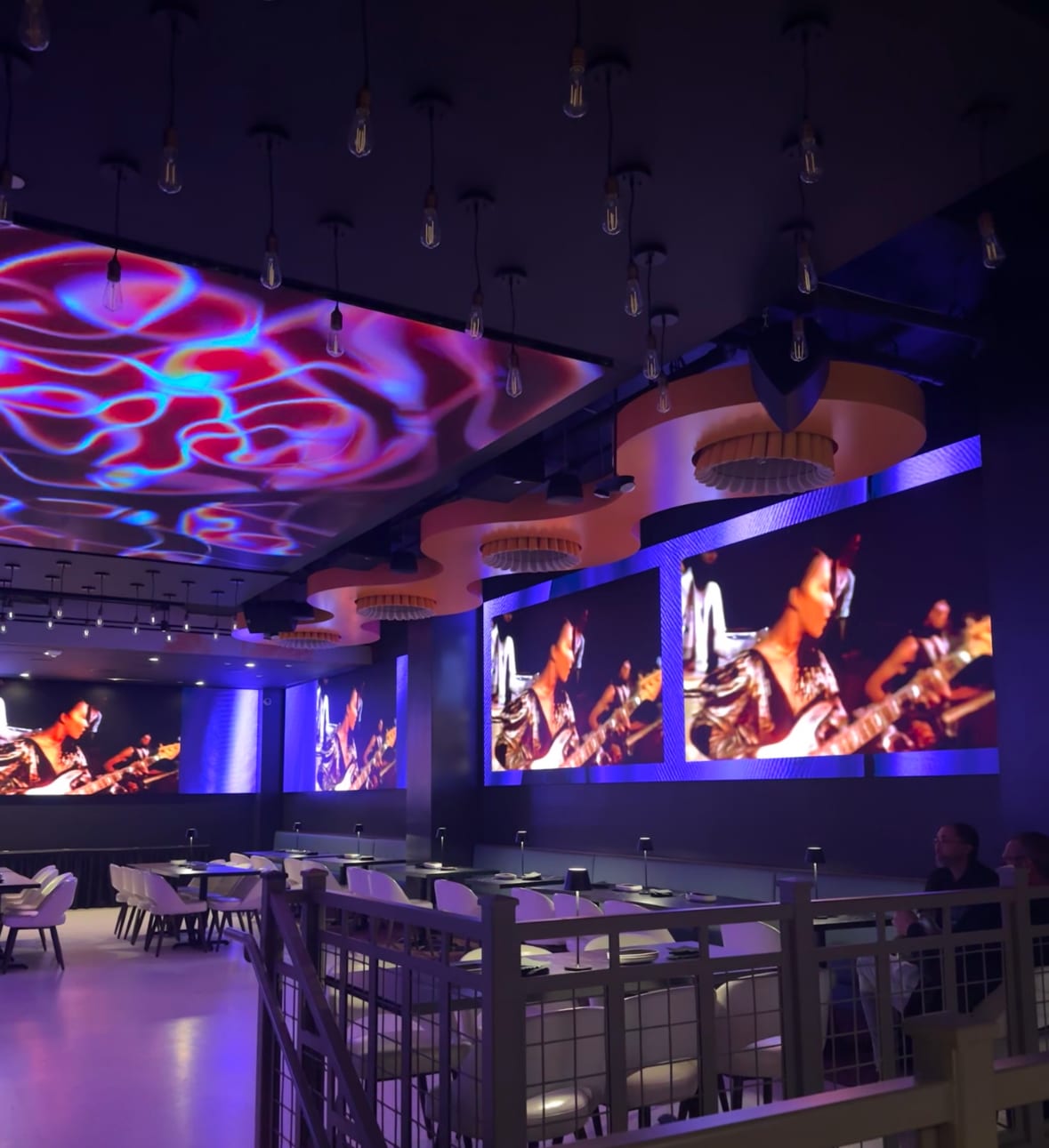 Planet Hollywood NYC interior with vibrant ceiling patterns, large LED screens displaying musicians, and modern dining tables.