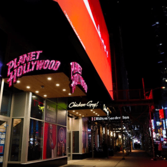 Night view of Planet Hollywood NYC storefront with illuminated signage, Chicken Guy! entrance, and Hilton Garden Inn nearby.